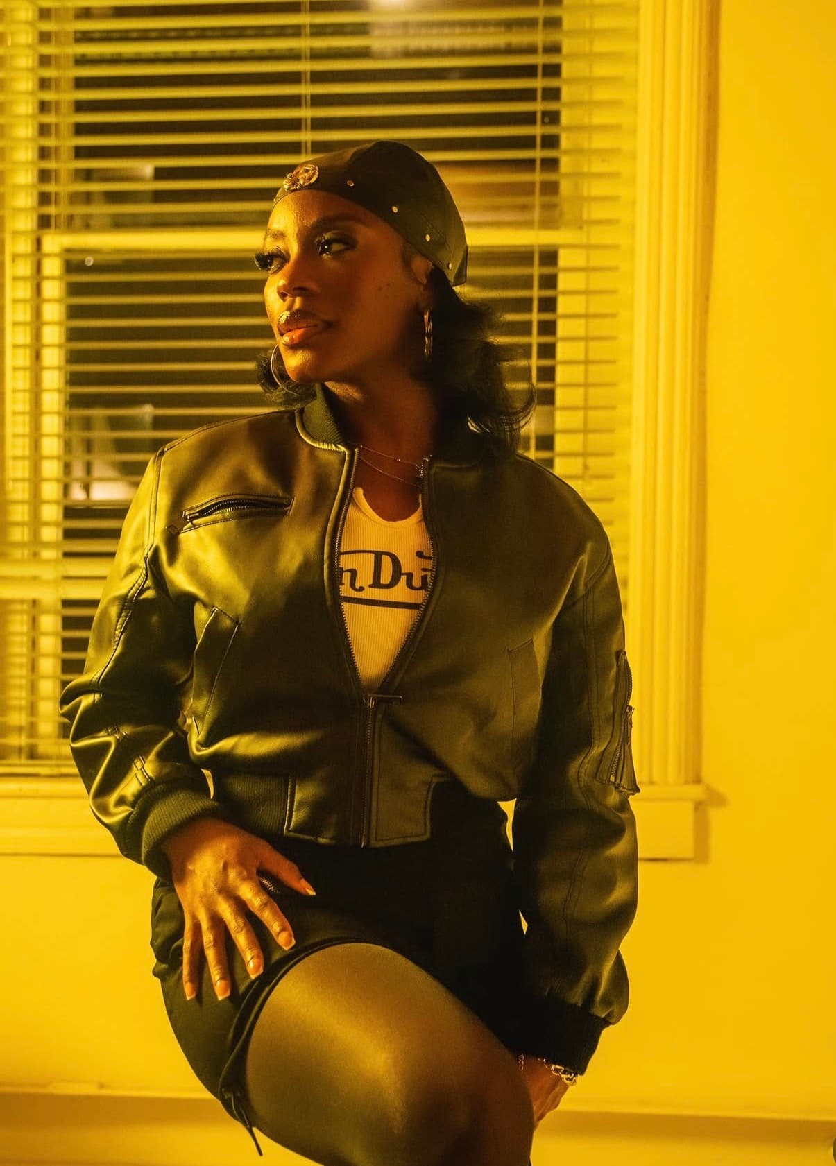 Tiyane Clarke seated by a window with blinds, wearing a black leather jacket, cap, and hoop earrings in warm golden light.