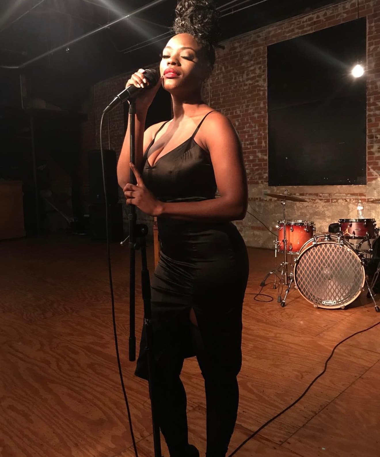 Tiyane Clarke singing into a microphone on a stage with exposed brick walls and a drum kit behind her.