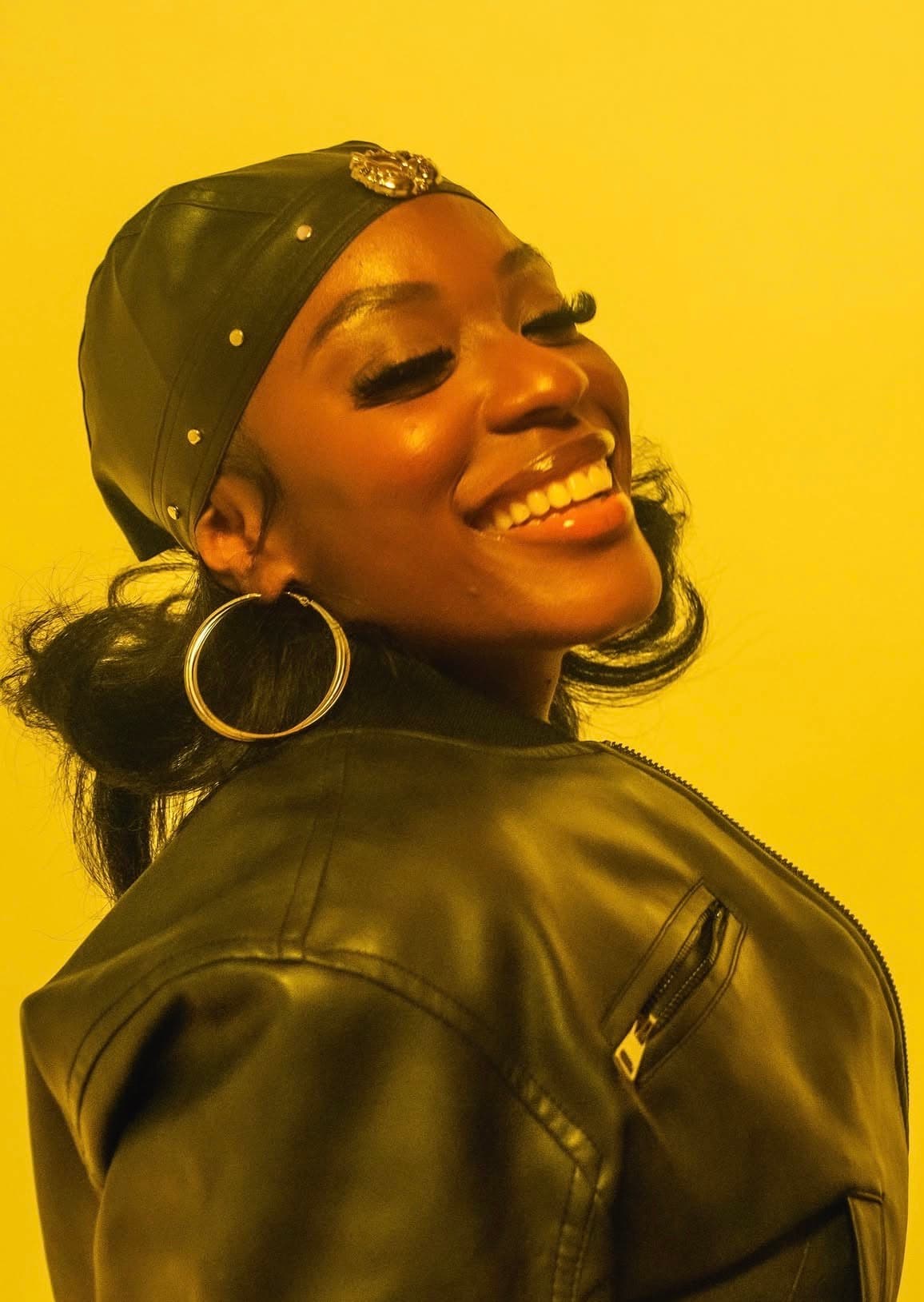 Tiyane Clarke smiling with her eyes closed, wearing a black leather cap, jacket, and large hoop earrings against a warm yellow backdrop.